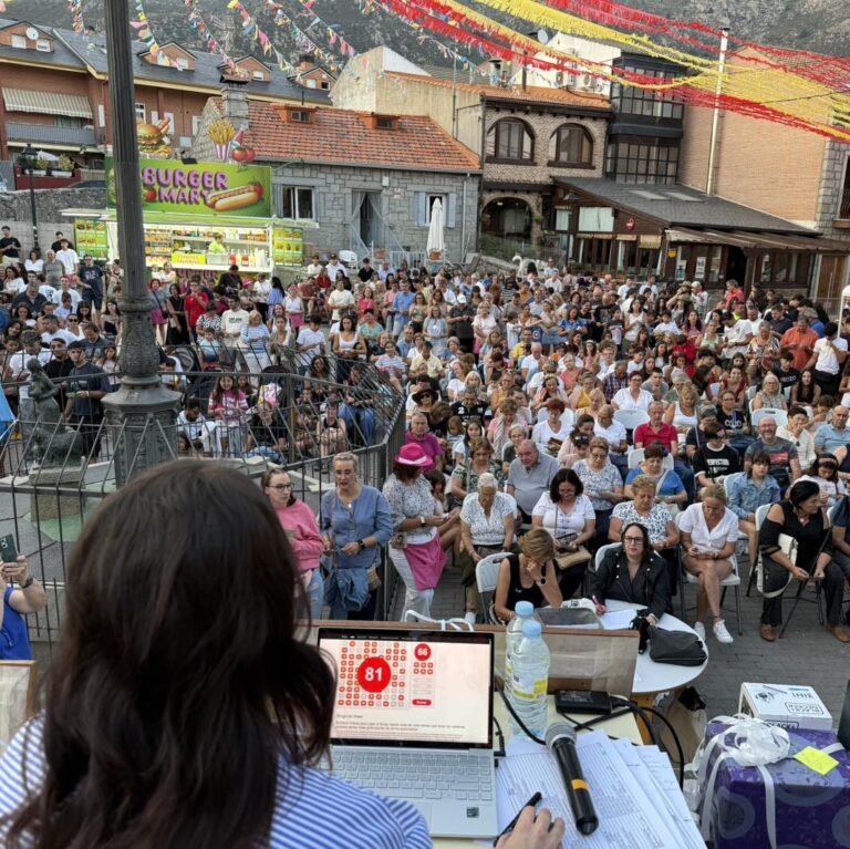 Una multitud de personas sentadas en una plaza, con una persona de espaldas en primer plano, operando un portátil que muestra un cartón de bingo en una pantalla, con banderines festivos al fondo.