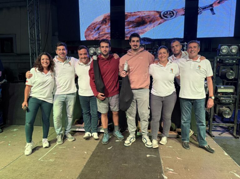 Un grupo de siete personas, incluyendo a miembros del Ayuntamiento, sonríen en un escenario con un trofeo en forma de jamón, en un evento festivo nocturno.