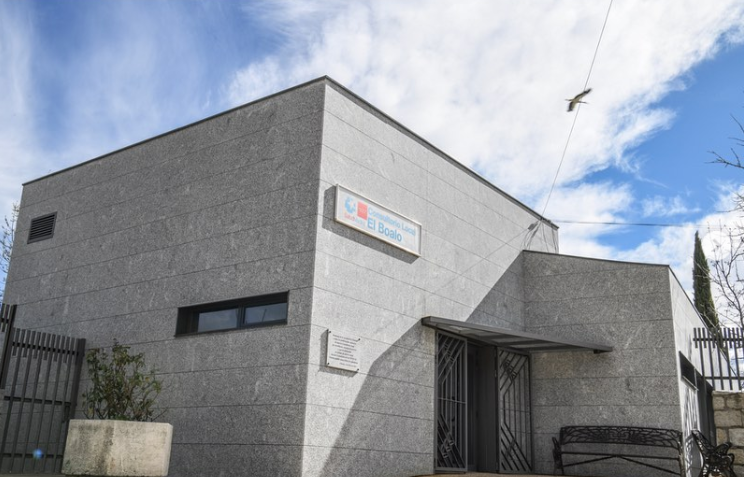 Fachada de un edificio de color beige bajo la luz del sol, con un banco de parque enfrente y un cartel de "Ayuntamiento El Boalo - Cerceda - Mataelpino Concejalía de Sanidad".