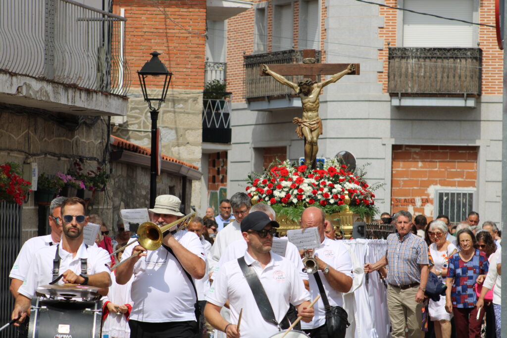 Una procesión religiosa en una calle de Cerceda, con una banda de música al frente y una figura de Cristo crucificado adornada con flores rojas y blancas, rodeada de personas.