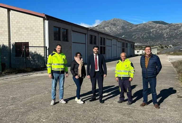 El alcalde Sergio Yunquera, de traje, posa con cuatro miembros de su equipo, algunos con chalecos de trabajo, frente a un gran edificio industrial con montañas al fondo.