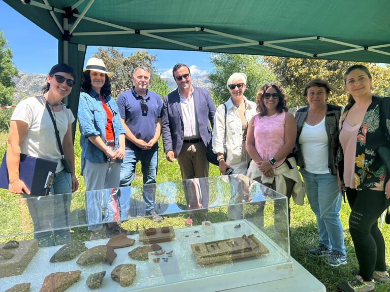 Un grupo de ocho personas sonrientes, incluyendo hombres y mujeres, posan alrededor de una vitrina de cristal que exhibe artefactos arqueológicos bajo una carpa verde al aire libre, con montañas al fondo.