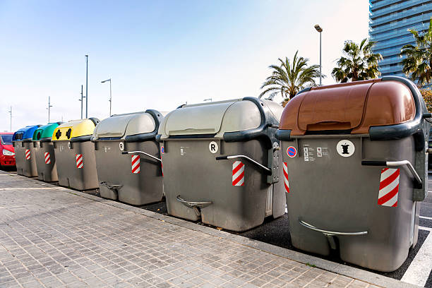 Una fila de contenedores de reciclaje de diferentes colores (rojo, verde, amarillo, gris, marrón) alineados en una calle urbana.