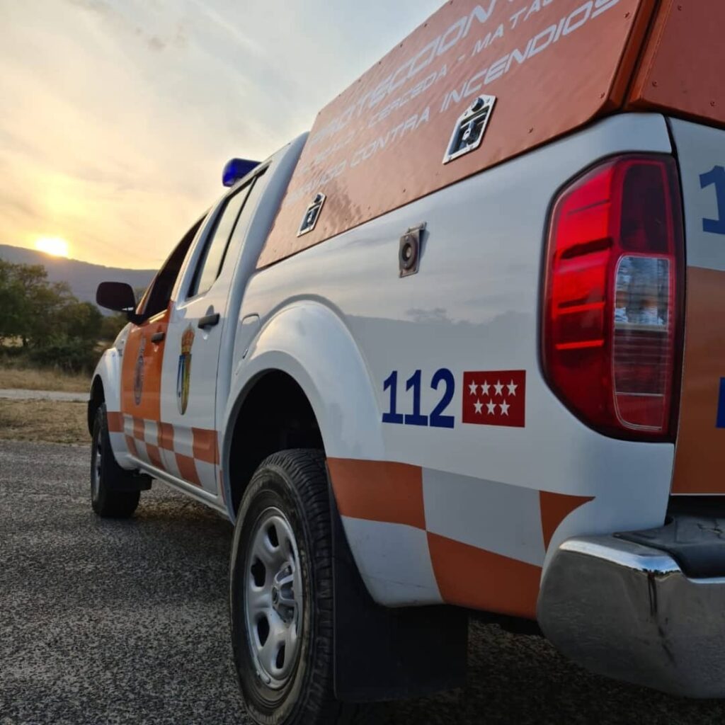 Parte trasera y lateral de un vehículo de Protección Civil blanco y naranja, con la numeración "112" y las estrellas de la Comunidad de Madrid, al atardecer en un paisaje rural.