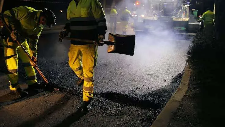 Dos trabajadores con chalecos reflectantes y herramientas, colocando asfalto caliente en una calle de noche, con maquinaria pesada y vapor al fondo.