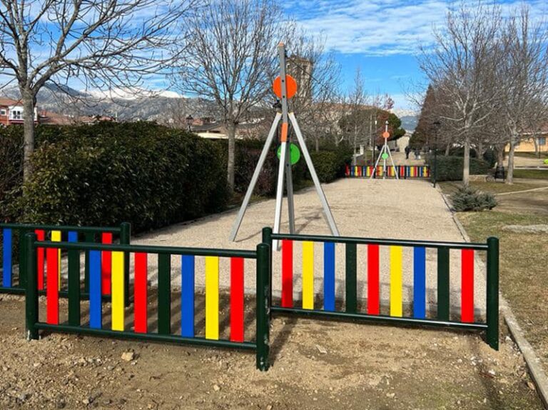 Un parque infantil con juegos de colores brillantes (rojo, amarillo, azul) y vallas de colores, rodeado de arbustos y árboles, con montañas nevadas al fondo.