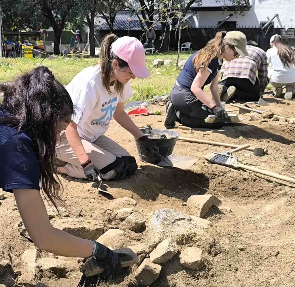 Cuatro mujeres jóvenes, algunas con gorras y guantes, arrodilladas en el suelo, realizando trabajos de excavación arqueológica con pequeñas herramientas y cubos en un yacimiento al aire libre.