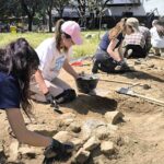 Cuatro mujeres jóvenes, algunas con gorras y guantes, arrodilladas en el suelo, realizando trabajos de excavación arqueológica con pequeñas herramientas y cubos en un yacimiento al aire libre.