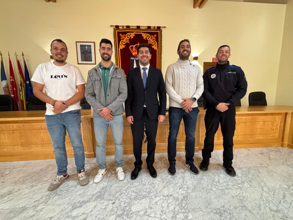 Cinco hombres, incluido el alcalde Sergio Yunquera y un agente de policía, posan sonrientes en un salón amplio con paneles de madera y banderas al fondo.