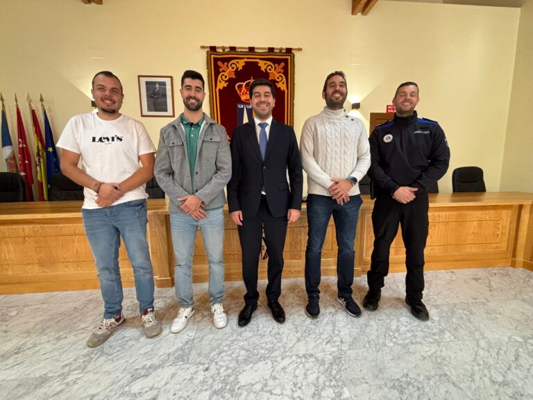 Cinco hombres, incluido el alcalde Sergio Yunquera y un agente de policía, posan sonrientes en un salón amplio con paneles de madera y banderas al fondo.