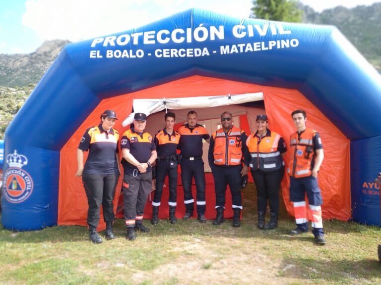 Un grupo de siete miembros de Protección Civil, con uniformes naranjas y azules, posando sonrientes frente a una gran carpa inflable de "Protección Civil El Boalo - Cerceda -Mataelpino".