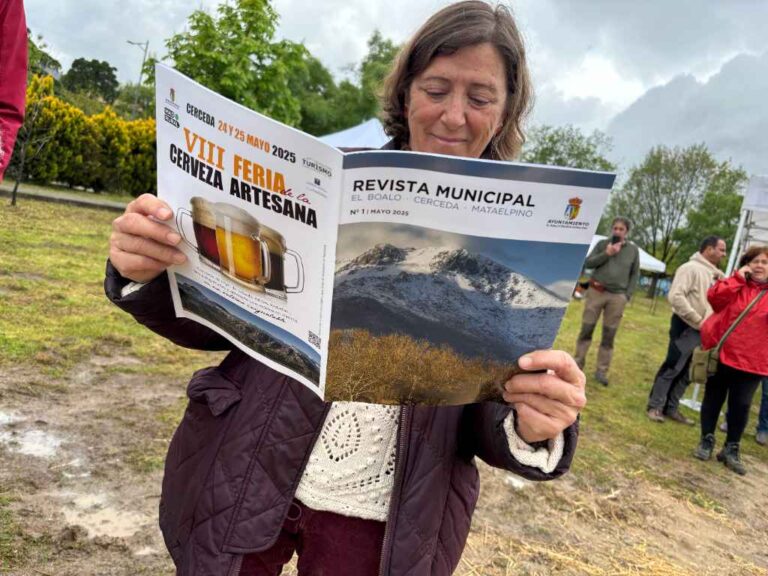 Una mujer de mediana edad leyendo una "REVISTA MUNICIPAL" abierta, que promociona la "VIII FERIA de la Cerveza Artesana" y muestra un paisaje montañoso.