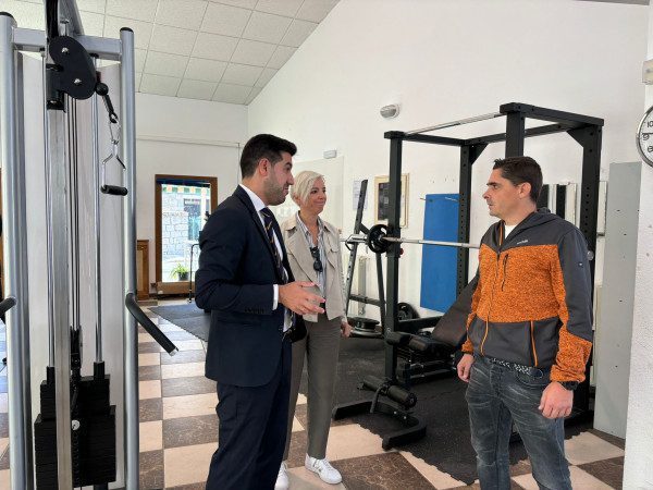 Interior del gimnasio con máquinas de pesas y tres personas conversando.