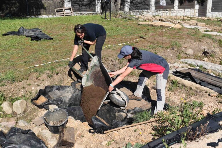 Dos personas, una con una pala y otra empujando una carretilla, trabajando en la remoción de tierra en un sitio de excavación arqueológica al aire libre.