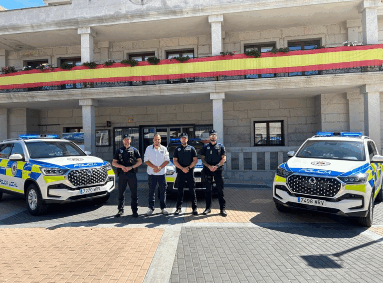 Cuatro hombres, incluido el alcalde Sergio Yunquera y agentes de policía, de pie frente a dos vehículos patrulla nuevos, con la fachada de un edificio adornada con una bandera española al fondo.