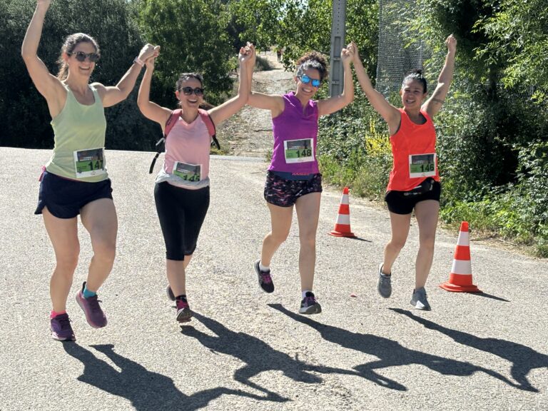 Cuatro mujeres corriendo y saltando con los brazos en alto, con dorsales y camisetas deportivas, en un camino rodeado de árboles en un día soleado.