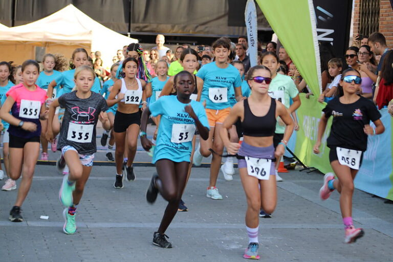 Un grupo de niños y niñas de diferentes edades corren en una carrera popular en la calle, con público observando al fondo.