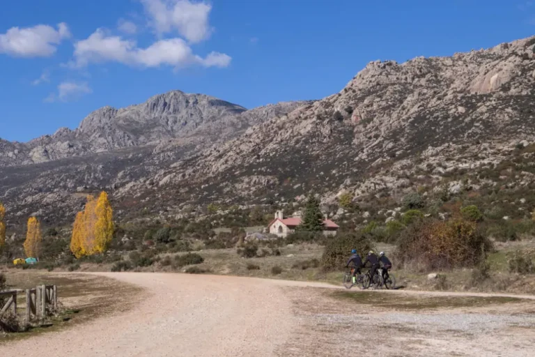 Grupo de ciclistas en un camino de tierra, con una pequeña ermita de piedra y montañas rocosas al fondo bajo un cielo azul con nubes blancas.