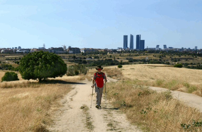 Un peregrino camina por un sendero de tierra en un paisaje seco, con el horizonte de la ciudad de Madrid y sus rascacielos visibles en la distancia.