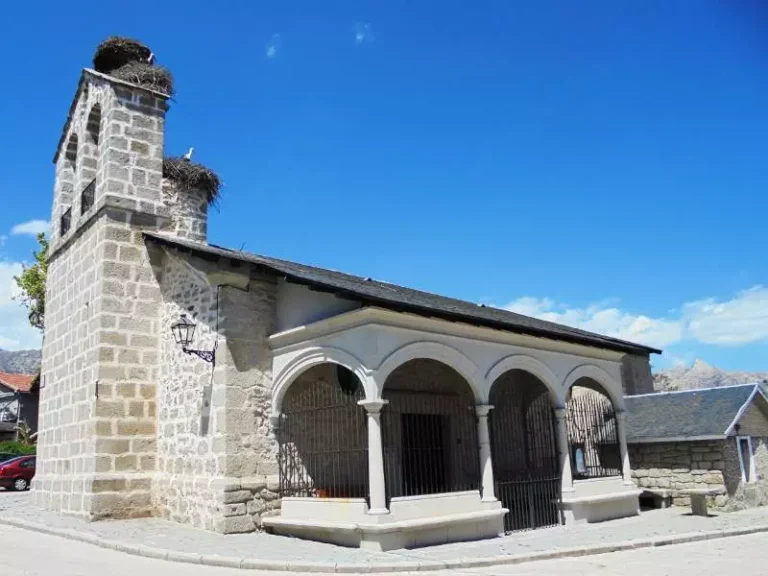 Fachada de piedra de la Iglesia de San Sebastián Mártir en El Boalo, con campanario y nidos de cigüeñas, bajo un cielo azul.
