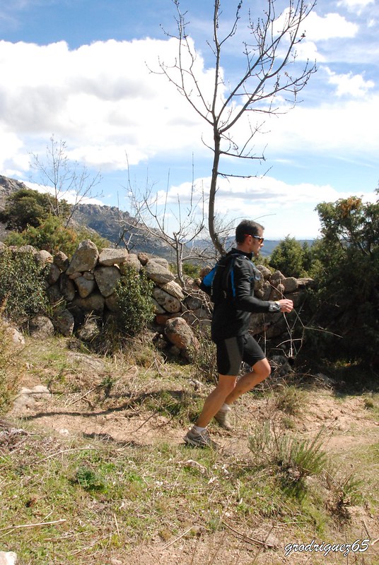 Un corredor de trail con gafas de sol y ropa deportiva oscura corre por un sendero de montaña rocoso con una pared de piedra.