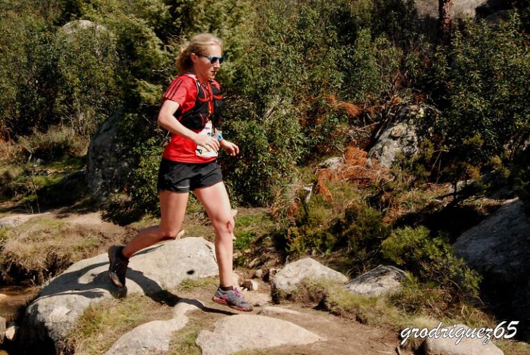 Una mujer con ropa deportiva roja y negra corre sobre rocas en un sendero de trail, saltando un pequeño arroyo.