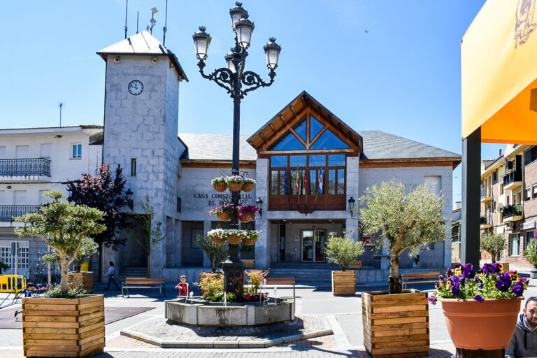 a fachada de la Casa Consistorial de El Boalo con una torre de reloj, una plaza ajardinada con bancos, farolas y fuentes, bajo un cielo azul brillante.