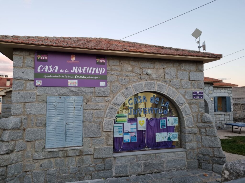 Fachada de piedra de la "Casa de la Juventud" con un letrero morado y ventanas cubiertas por contraventanas, en un entorno con árboles y cielo al atardecer.