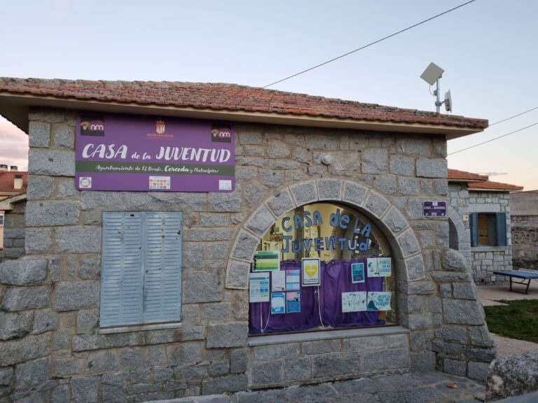 Fachada de piedra de la "Casa de la Juventud" con un letrero morado y ventanas cubiertas por contraventanas, en un entorno con árboles y cielo al atardecer.
