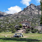 Una pequeña ermita de piedra con tejado rojo, ubicada en una colina rocosa con vegetación, al pie de una gran montaña rocosa, bajo un cielo azul con nubes.
